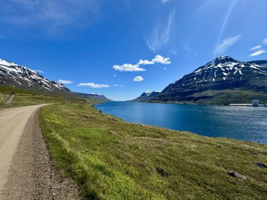 Güneşli bir günde, İzlanda fiyordunun mavi suyunun yanındaki asfaltsız yol karlı dağlarla çevrilidir.