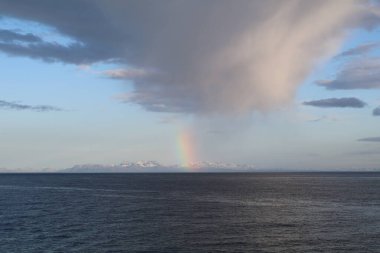 the ocean with stormy clouds in the distance, a blue sky, and a rainbow emerging from the clouds into the ocean clipart