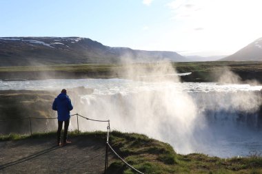 silhouette of a single man in front of the impressive waterfall Godafoss at sunset with the spray of the fall being lit by the sun clipart