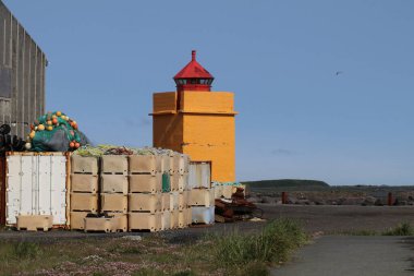 A vibrant yellow lighthouse with a red top stands against a clear blue sky in a coastal fishing area with in the foreground stacks of weathered storage crates, fishing nets, and buoys. clipart