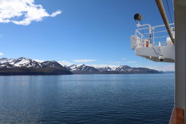 Sakin bir fiyorda bakan bir yolcu gemisinden çarpıcı bir manzara, yüksek dağlar ve açık mavi gökyüzünün altında karla kaplı tepeler.
