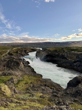 Volkanik bir arazide uzanan vahşi Skalfandafljot nehri. Uzaktaki Godafosss şelalesi altın saatin yumuşak ışığında dökülüyor.