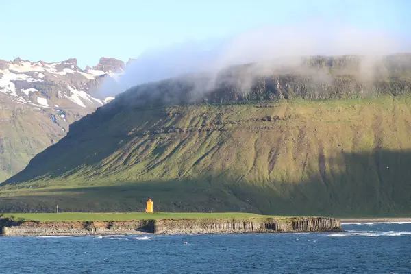 Sisli dağların altında bazalt sütunları olan engebeli bir sahil şeridinde uzak İzlanda deniz feneri.