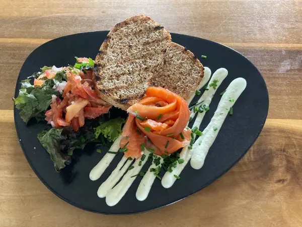 Tütsülenmiş somon balığının üst görüntüsü ve salata ile servis edilen tam tahıllı ekmek ve ahşap bir yüzey üzerinde siyah bir tabakta kaplanmış soğanlı kremalı sos..