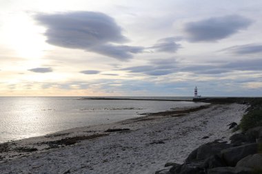 Sakin bir okyanusu, boş kumlu bir plajı ve uzakta bir deniz feneri silueti olan bir deniz pelerini. Bu bulutlu gökyüzünün altında, güneş doğuyor ve ufukta mavi gökyüzü.