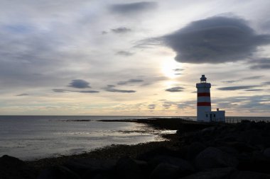 Alacakaranlıkta bulutlu bir gökyüzünün altında kırmızı ve beyaz bir deniz feneri olan kayalık İzlanda kıyısı