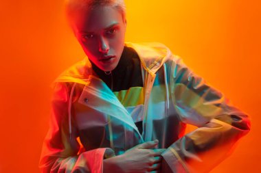 Androgynous kadın modeli kameraya bakıyor ve şeffaf fütürist yağmurluklara sarılıyor parlak neon ışığı altında turuncu arka planda