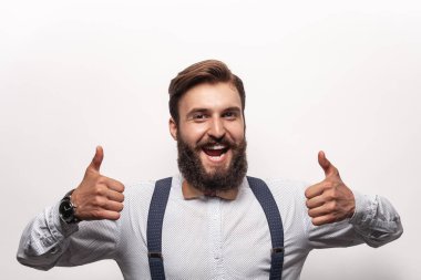 Şık giyinmiş neşeli sakallı erkek hippi gülümseyerek kameraya bakıyor ve gri arka plana başparmak işareti yapıyor.