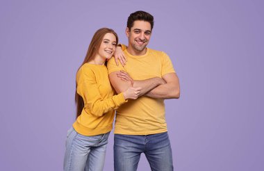Sarı tişörtlü, kot pantolonlu, gülümseyen ve kameraya bakan genç, kızıl saçlı kadın, kollarını mor arka planda çapraz bağlamış erkek arkadaşının omzuna yaslanırken,