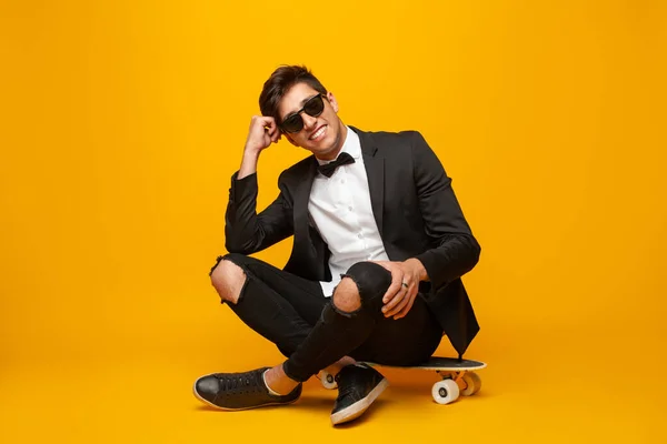 黄色の背景を背景にスケートボードに座っている間に手にもたれ 笑顔でカメラを見て スタイリッシュな服やサングラスで全身うれしい若い男性 — ストック写真