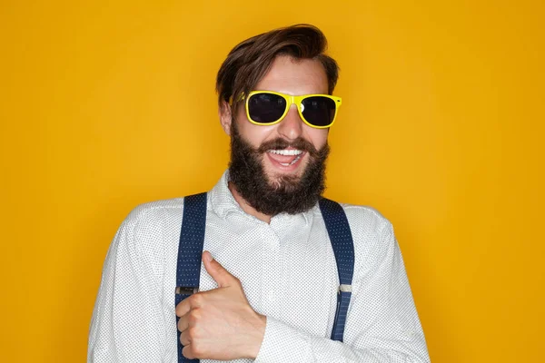 快乐的大胡子男人 身穿时髦的衣服 戴着太阳镜 微笑着看着相机 用手指在黄色的背景下摆出姿势 — 图库照片