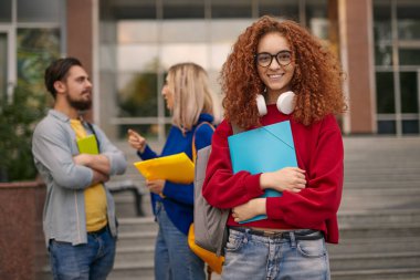 Kıvırcık saçlı, günlük kıyafetler ve gözlüklü, gülümseyen ve kameraya bakan üniversite binasının yanında elinde klasörle arkadaşlarının yanında duran pozitif bir genç kız öğrenci.