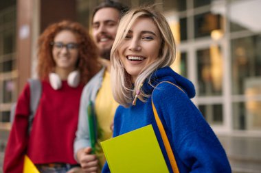 Cheerful young female student in blue hoodie with blond hair looking at camera and smiling while standing near friends during university admission process clipart