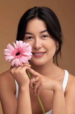 Mutlu Asyalı kadın, siyah saçlı taze Gerbera çiçeğiyle yüzüne dokunuyor ve kahverengi arka planda gülümseyerek kameraya bakıyor.