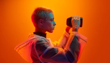 Yağmurluk giyen, kısa saçlı, VR gözlüklerini inceleyen turuncu arka planda neon ışığı altında duran genç bir kadının yan görüntüsü.