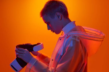 Fütürist yağmurluklu genç çift cinsiyetli modelin yan görünüşü. Kısa saçlı, turuncu arka planda neon ışığı altında VR kulaklığı takıyor.