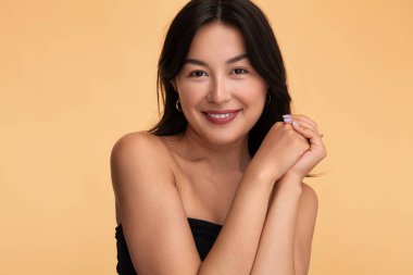 Mutlu Asyalı bir kadın, çıplak omuzlarıyla elini sıkar ve bej arka planda gülümseyerek kameraya bakar.