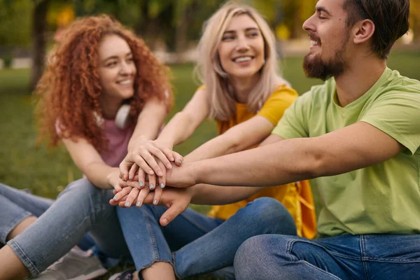 正の若い男性と女性の友人でカジュアル服笑顔とスタッキング手で一緒に座っている間草の芝生の上で公園 — ストック写真