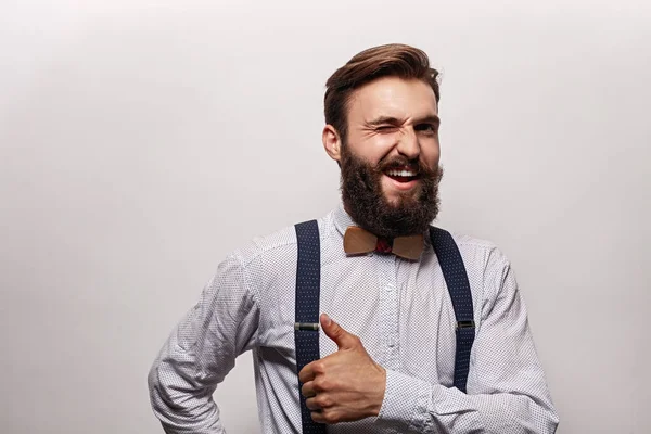 身穿时髦服装的快乐大胡子男人对着相机眨眼 并在灰色背景下显示出大拇指的姿势 — 图库照片