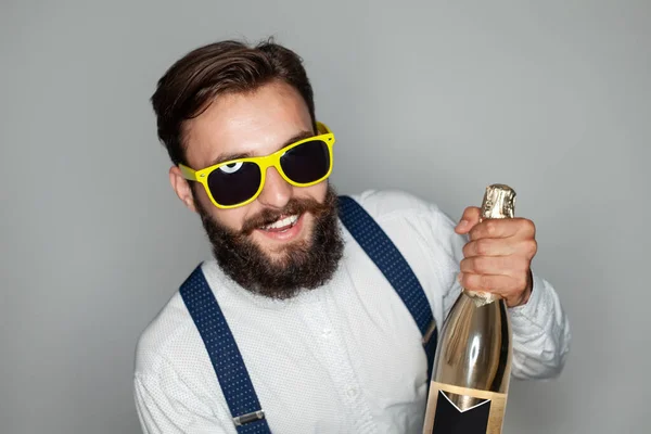 スタイリッシュな服やサングラスで幸せな髭を生やした男性ヒップスターシャンパンのボトルを示し 灰色の背景を背景にパーティー中に笑顔でカメラを見て — ストック写真