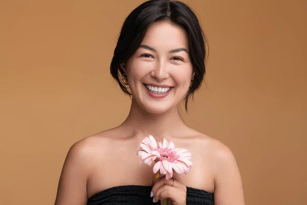 若いですアジアの女性裸の肩と黒髪保持新鮮なピンクのガーベラの花と茶色の背景に笑顔でカメラを見て — ストック写真