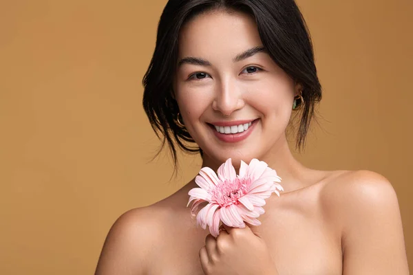积极的年轻亚洲女性 有勇敢的肩膀 触碰下巴 有粉红色的德国花 在棕色背景下微笑着看着相机 — 图库照片