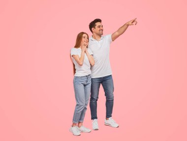 Pembe arka planda yan yana dururken, tüm vücudunu mutlu eden, günlük kıyafetlerle gülümseyen ve şaşırmış kız arkadaşına işaret eden genç adam.