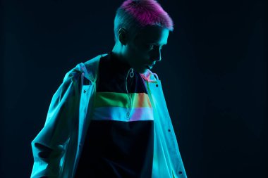 Genç çift cinsiyetli, şık, fütüristik elbiseli, kısa saçlı bir kadın siyah arka planda renkli neon ışığın altında dikilirken gözlerini kaçırıyor.