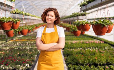 Beyaz tişörtlü ve sarı önlüklü mutlu bir kadın çiftlikteki seranın yanında dikilirken kameraya gülümseyerek bakıyor..