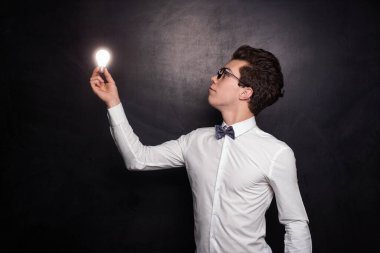 Siyah saçlı, beyaz gömlekli, papyonlu ve gözlüklü genç bir erkek öğrenci elinde ampulle siyah arka planda duruyor.