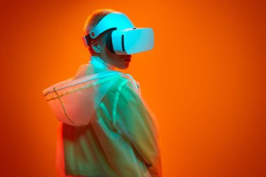 Fütürist yağmurluk ve VR kulaklık takan genç bir kadın, turuncu arka plana karşı neon ışığı altında sanal gerçeklik deneyimi yaşarken omzunun üzerinden başka tarafa bakıyor.