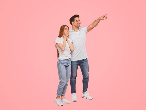 Full body happy young man in casual clothes smiling and pointing away for amazed girlfriend while standing against pink background together