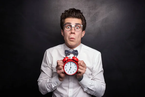Scared young man in white shirt with bow tie and glasses, showing alarm clock and looking at camera while being late to studies against black background