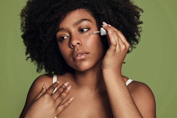非洲裔美国女性 卷发在脸颊上滴下保湿血清 在绿色背景的护肤过程中 眼睛移开 — 图库照片