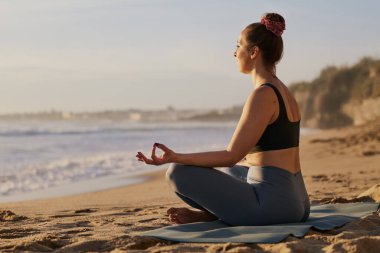 Aktif kıyafetli genç bir kadın. Gyan Mudra 'yı işaret ediyor ve Lotus' ta meditasyon yapıyor. Yoga sırasında deniz kenarındaki kumsalda poz veriyor.