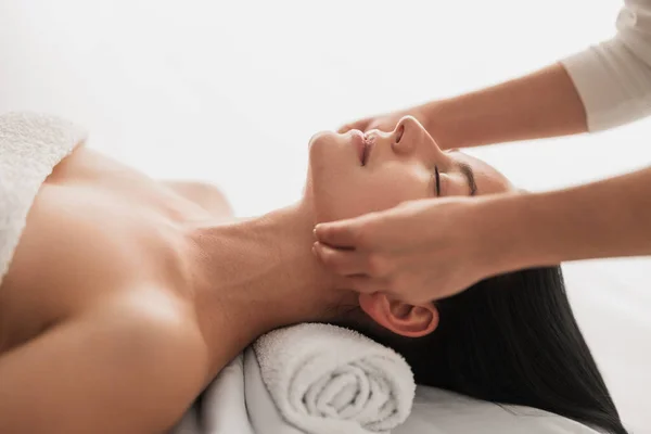 Side view of crop unrecognizable cosmetician doing facial massage to relaxed young female, customer with closed eyes and bare shoulders lying on table during beauty procedure in light salon