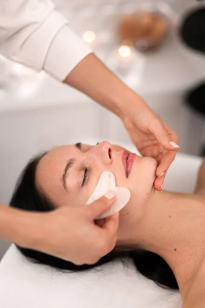 From above of crop anonymous cosmetician doing face massage with gua sha tool to peaceful young female client with dark hair, lying on couch with closed eyes in salon