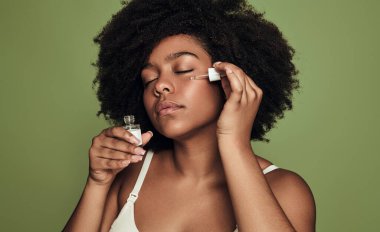 Koyu Afro saçlı ve kapalı gözlü genç siyah kadın milenyumu, yeşil arka plana karşı cilt rutini sırasında pipette ile yüzüne hyaluronik asit serumu uyguluyor.