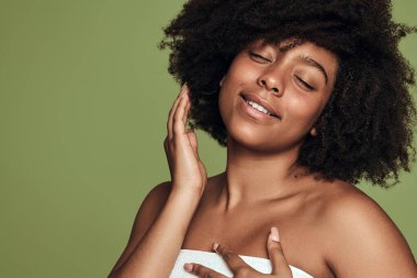 Mükemmel ciltli, çıplak omuzlu, Afro saçlara dokunan ve yeşil arka planda gülümseyen genç, zevkli bir etnik kadın modeli.