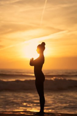 Silhouette of woman meditating in Mountain pose with prayer hands against waving sea and bright sunset sky during yoga session in evening on beach