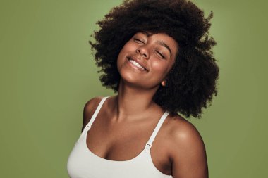 Delighted young black female model with perfect skin and dark Afro hair in white bra, smiling happily with closed eyes against green background in studio