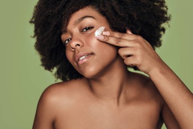 Optimistic African American female with curly hair smiling and looking at camera while applying moisturizing cream over green background in studio