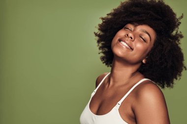Side view of confident happy young black female model with dark Afro hair in white bra smiling with closed eyes against green background