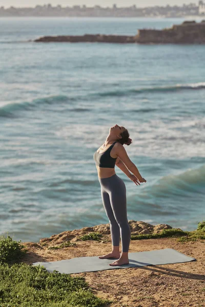 Side view full body of young barefooted woman in leggings and top stretching arms and back, while standing on mat during yoga session on rocky shore near wavy ocean