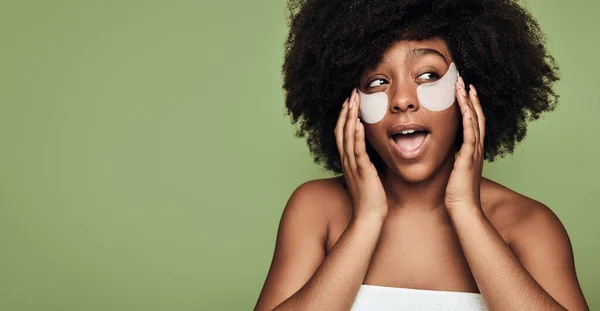 stock image Positive confident young black female millennial with Afro hair, applying hydrating collagen patches on face and looking away with opened mouth against green background