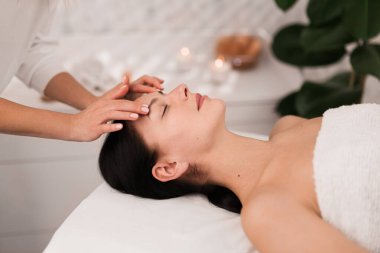 Crop anonymous female cosmetician doing face massage to peaceful young client with dark hair, lying on couch with closed eyes during skincare procedure in modern beauty salon