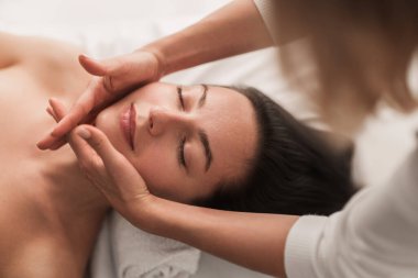 High angle of young female lying on bed with closed eyes while therapist makes a relaxing face massage procedure at spa