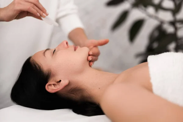 Side view of young relaxed female client with long dark hair and bare shoulders, lying on table while crop unrecognizable cosmetician doing face massage with gua sha