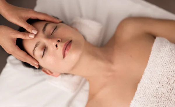 Top view of young female with bare shoulders lying on bed while her head is being massaged by professional face masseur at spa