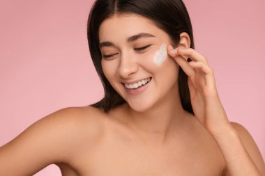 Crop young delighted female millennial with long dark hair and bare shoulders, smiling brightly and looking away while applying skincare cream on face against pink background
